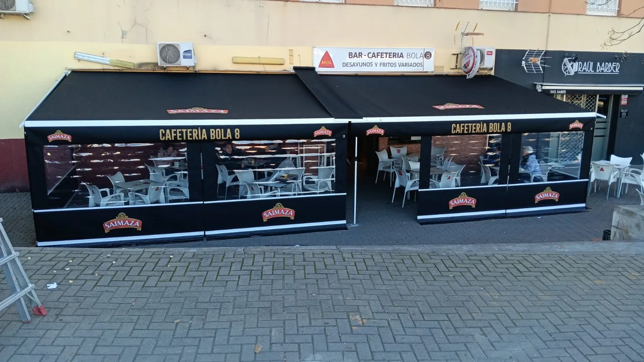 Terraza de la cafetería Bola 8 en Alcalá de Guadaíra, con lonas negras rotuladas Saimaza y ventanas transparentes